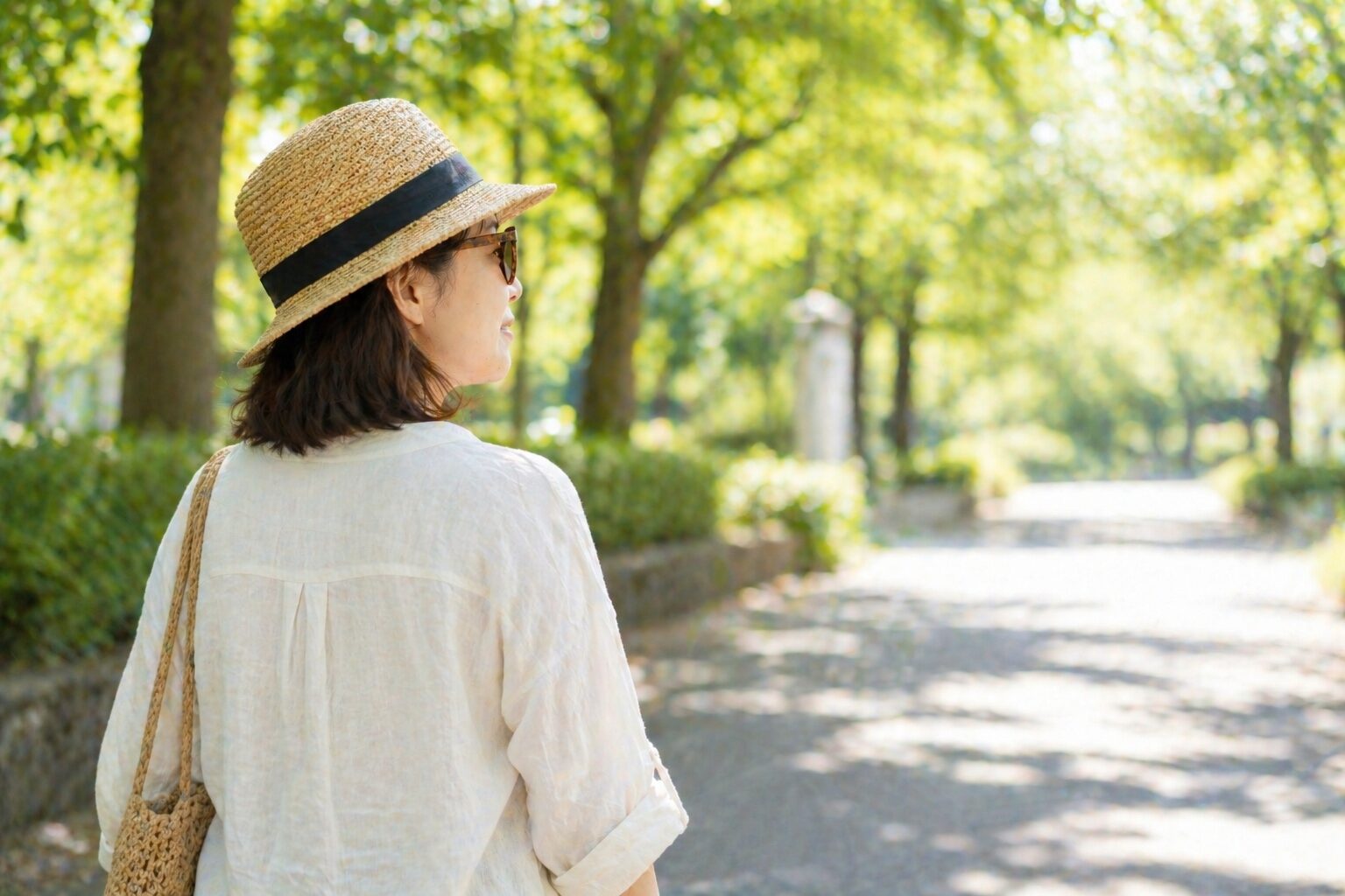 木漏れ日の公園道を、帽子とサングラスを着けて歩く女性の後ろ姿。紫外線から目を守りながら外出を楽しむイメージ